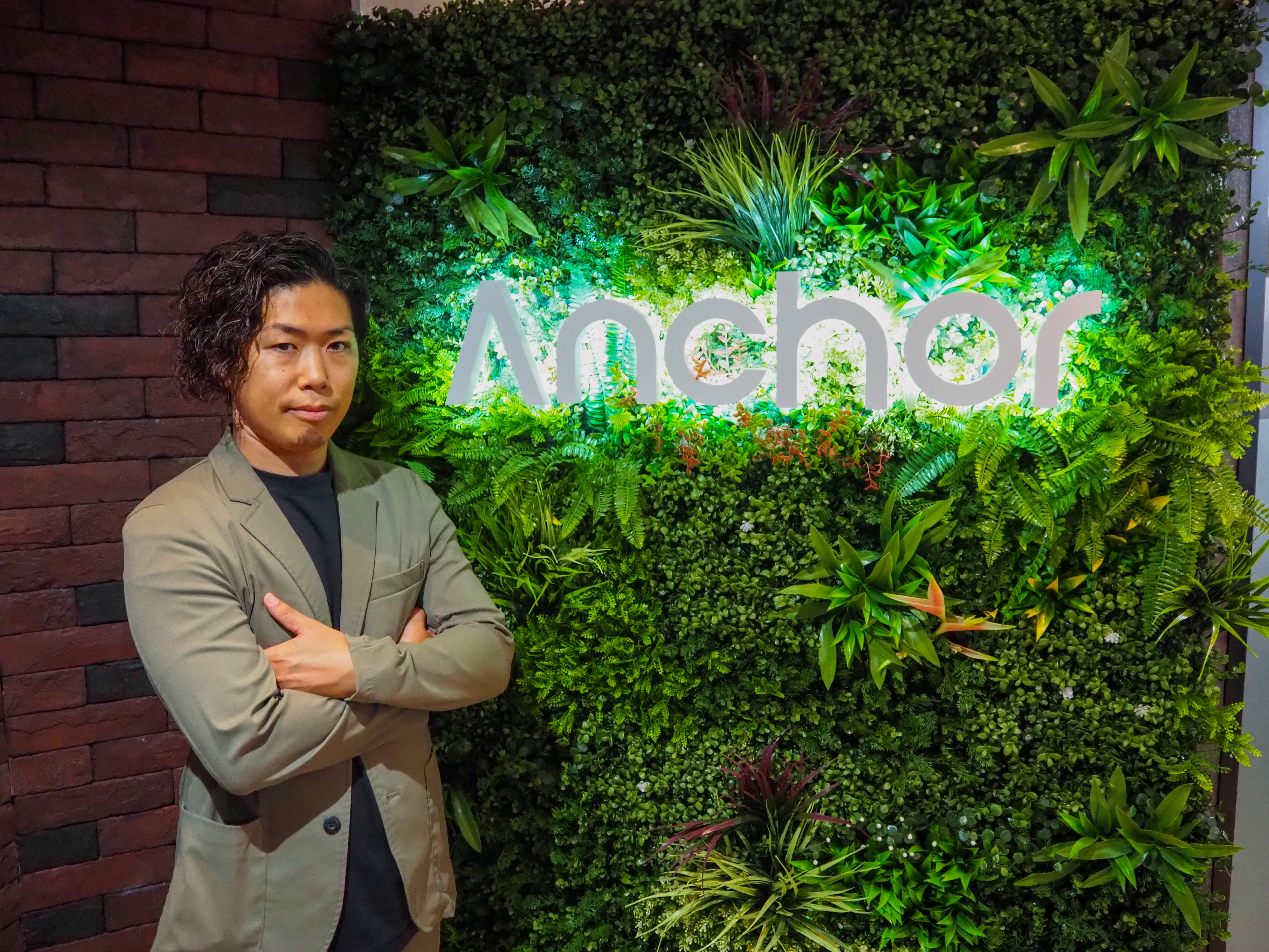 アンカー株式会社のロゴが入ったグリーンウォールの前に立つ、執行役員の北 哲哉さん