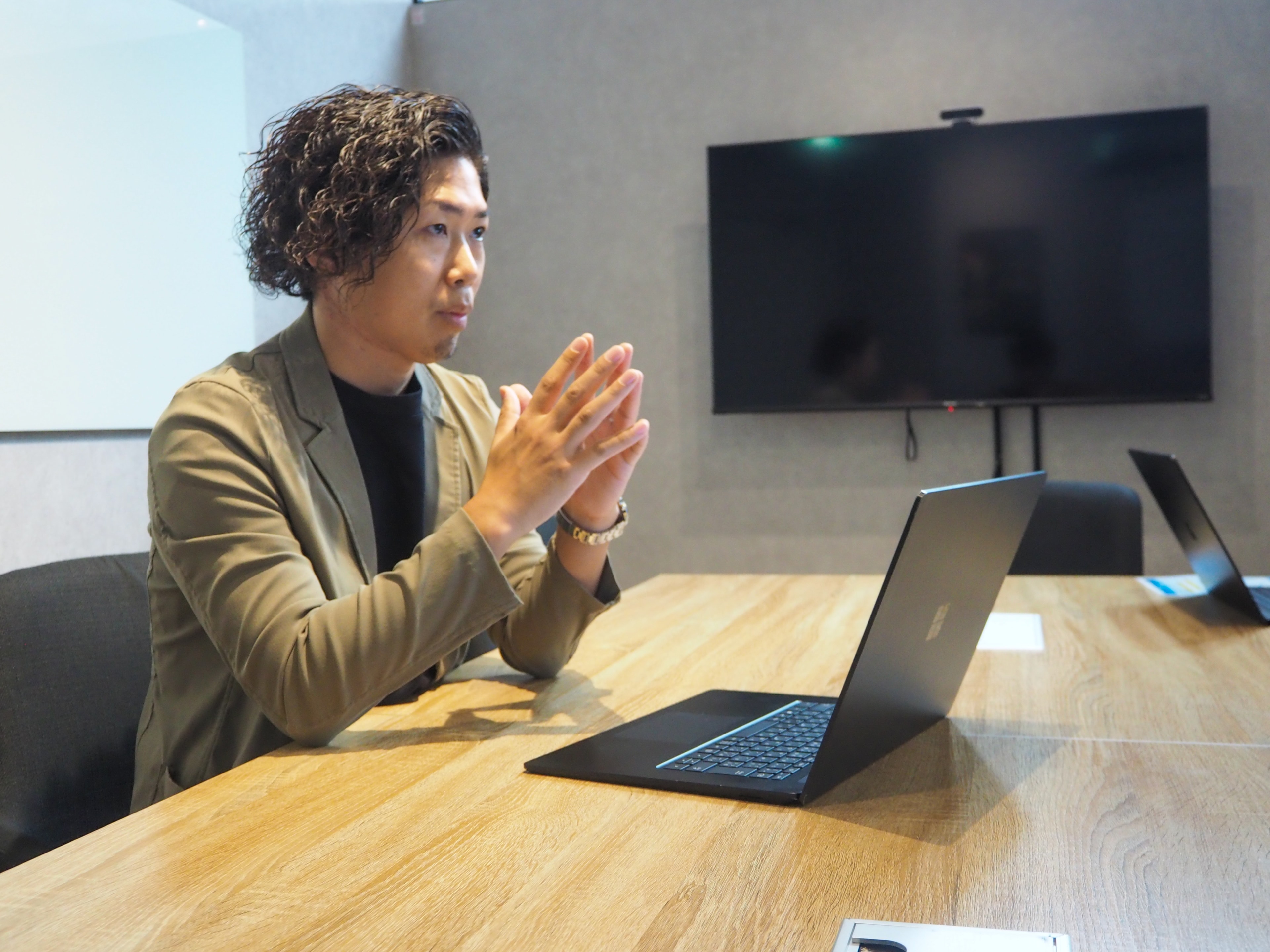 大型モニターのある会議室で、手を合わせながら成果を語る北さん